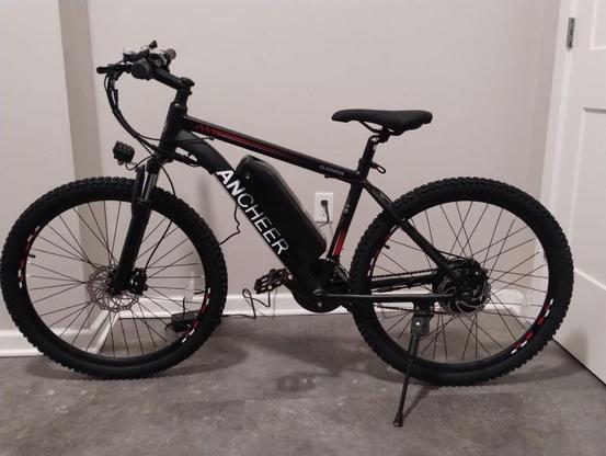 Full size black mountain bike (with red and white highlights) with a large battery on the bottom frame, plugged into the wall behind it with a charger