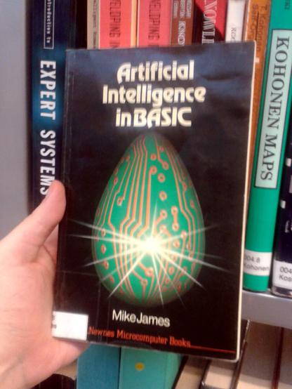 A person holds a book titled "Artificial Intelligence in BASIC" by Mike James. The cover features a green circuit-patterned egg with a bright starburst. Bookshelves in the background. (Yes, this caption was made by AI, and I'll present it unedited, because it just happens to be accurate. I usually have to edit these a lot.)
