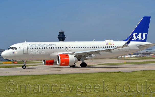 Side view of a white, twin engined jet airliner taxiing from right to left along a grey runway.
The plane is mostly white, with silver "Scandinavian Airlines" titles above and below the forward cabin windows, and a dark blue tail with white "SAS" titles.
The engine pods under the wings are bright orange at the front, with white "Scandinavian" titles, and a white rear section.
Green grass fills the foreground, with a tall black tower looming over the plane in the background, under blue-grey skies.