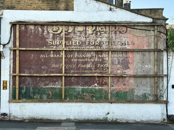 An exterior wall with a wooden lattice structure for mounting a billboard visible. In the absence of the billboard, a fading hand-painted sign advertising Boyd Pianos is visible. Photo via History of Stoke Newington.