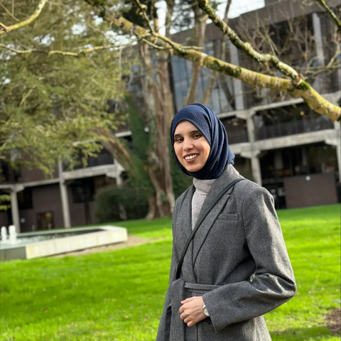 Photograph of a woman in a grey coat stood outdoors with large trees and a large glass-fronted building behind her. She is smiling at the camera