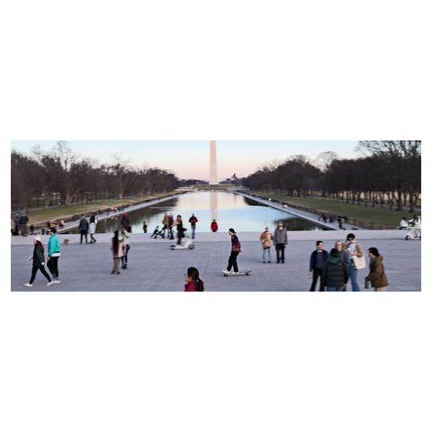 View of the pool and the Washington monument with people around blurred and one person passing by on a skate