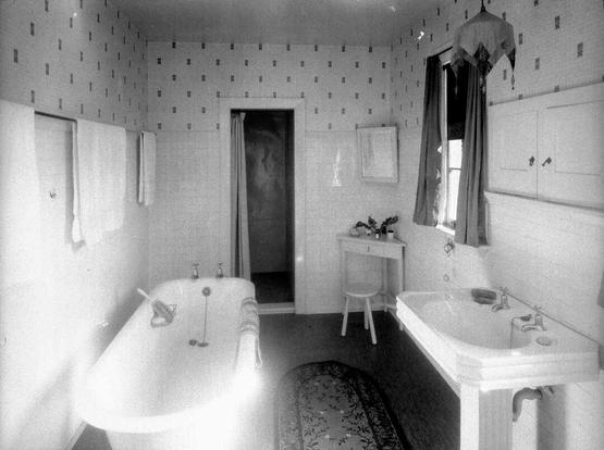 Black-and-white photo: Bathroom interior. ca. 1920. Photographer, Steffano Webb. Description: View of a new bathroom. Visible are the bath, wash basin, corner table, towels and towel rail, mirror, light shade and curtains. The lower part of the walls are covered with plain white tiles; the upper walls are papered. It represents a state-of-the-art bathroom for its time. Citation: Webb, Steffano, 1880-1967: Collection of negatives. Ref: 1/1-003914-F. Alexander Turnbull Library, Wellington, New Zealand. https://natlib.govt.nz/records/22883310