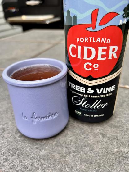 A close-up image of a glass of cider in a light purple ceramic cup next to a bottle of Portland Cider Co. Tree & Vine. The background features a textured surface and hints of outdoor elements.