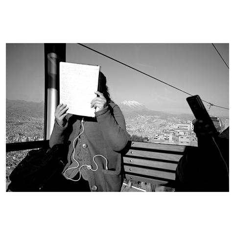 Photo of a person reading her notebook inside a cablecar cabin, with the snowy mountain Illimani in the background. Taken in La Paz, Bolivia.