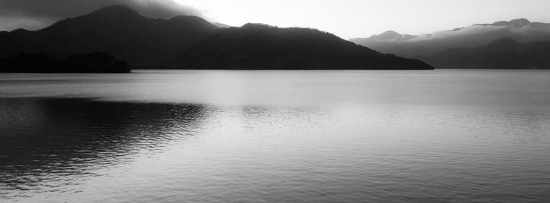The image depicts a serene landscape with a calm body of water in the foreground. In the background, there are silhouettes of hills or mountains, with some clouds hovering above them. The scene is in black and white, creating a tranquil and timeless atmosphere.