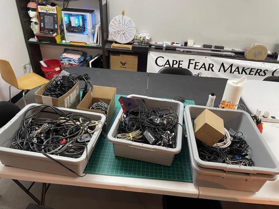 Picture of multiple large plastic bins and cardboard boxes filled with a mess of random cables. Every thing is sitting on a table.
