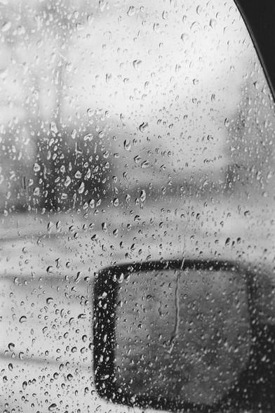 Close-up of a rainy car window covered in water droplets, with a blurry side mirror and indistinct outdoor scenery visible through the glass. The image is black and white, creating a moody, introspective atmosphere.