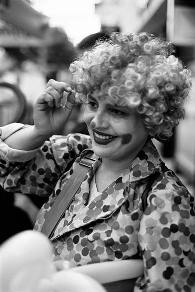 A black and white close-up shot shows a clown at a street art festival. The clown has a wide smile, revealing their teeth, and their gaze is directed slightly off to the right. They have dark, painted eyebrows, and their lips are painted a dark shade, extending in a wide, exaggerated smile typical of clown makeup. Round, dark smudges are visible on their cheeks.

The clown is wearing a curly, light-colored wig that is voluminous and frames their face. Their attire consists of a jacket with large, irregular polka dots in varying shades of gray and white. A strap, possibly part of their costume or an accessory, goes over their shoulder. One hand is raised and appears to be adjusting the wig near their forehead. The background is blurred, suggesting an outdoor setting with other people and possibly elements of the festival.