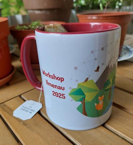 A tea pot stands on a wood-plank outdoor or garden  table with some brown plant pots in the background. The mug is white on the outside, with the printed logo of YoMos half visible on the right and the text "workshop 2025 Ilmenau" written in dark red on the right, next to the handle. Both handle and the inside of the mug are colored dark red.