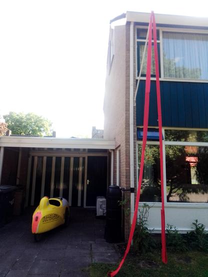 Velomobile parked outside a house in the Netherlands with a long red line of knitted scarf dangling from the flagpole.