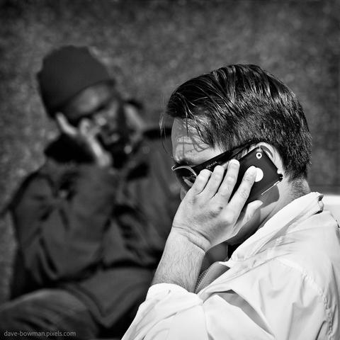 A photograph of a street scene in San Francisco captures a quiet moment shared by two men in contrasting circumstances. In the foreground, a well-dressed man wearing designer sunglasses talks intently on his phone, his posture focused. In the background, another man sits with his head resting in his hand, echoing the same gesture but who appears to be sleeping. The composition subtly highlights the differing realities that exist side by side in the city, offering a layered view of urban life.