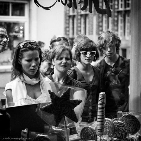 A photograph of a group of people standing outside a sweet shop in Bruges, Belgium, taken in 2008. They are gathered closely, leaning toward the shop window, their faces and postures focused with quiet curiosity. The monochrome tones highlight the contrast between the figures and the shopfront, drawing attention to the subtle expressions and body language as they look inside.