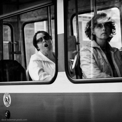 A photograph of two women on a passing bus, taken from the street as it goes by. One woman in the background sits mid-yawn, her mouth wide open in a perfectly timed, unguarded moment. Near the window, a button labeled “Open” adds a layer of quiet humour, aligning with the woman's actions. An ordinary moment is made memorable by its timing and subtle visual irony.