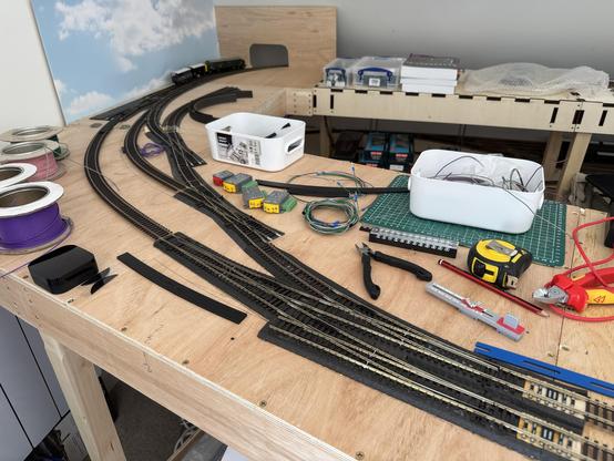 A model railroad setup featuring tracks on wooden baseboards, with various tools and wires scattered around. There are spools of wire, small model train cars, and a ruler. A white container holds additional wires, with a green cutting mat underneath.