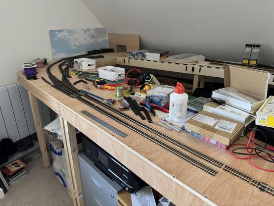 A cluttered model railway layout under construction. It features various tools, electrical components, rolls of wiring, and railway tracks arranged in curves. Boxes containing model accessories and supplies are visible, alongside a printer and other materials.