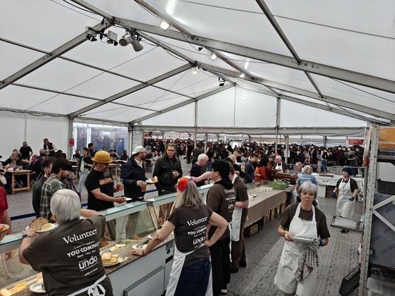A large tent with a lot of people. There are a number of fridges containing cheese and pies