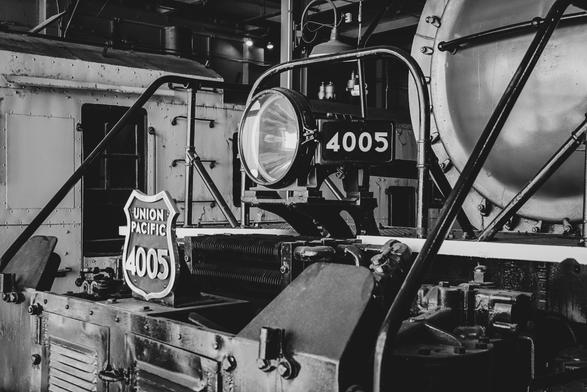 The image is a black and white photograph of a steam locomotive, specifically focusing on the front section. The locomotive is marked with the number "4005" and the name "Union Pacific" on a sign. The headlight is prominently displayed, mounted on a metal frame, and is circular with a large glass lens. The metal body of the locomotive is visible, showing rivets and a weathered texture, indicative of its age and use. The background includes parts of the locomotive's structure, such as a large cylindrical boiler and a metal railing. The image captures the intricate details of the locomotive's design and construction, emphasizing its historical significance.

Provided by @altbot, generated privately and locally using Ovis2-8B