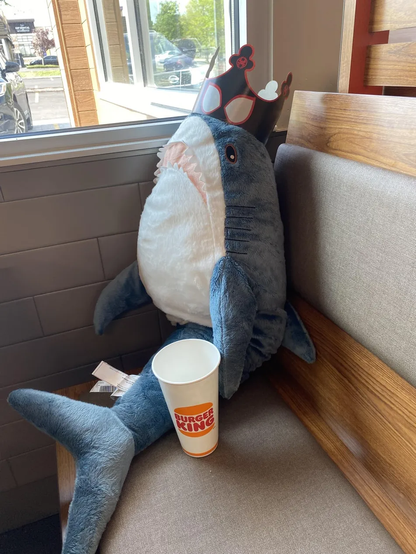 A plush BLÅHAJ shark sits in a Burger King booth by a window. The blue and white stuffed shark is wearing a paper crown with red trim and white eyeholes. The BLÅHAJ is positioned upright against the beige booth seating, with its fins spread out and a Burger King drink cup placed next to it on the bench. Sunlight streams in through the window, illuminating this whimsical scene of the shark enjoying a fast food outing