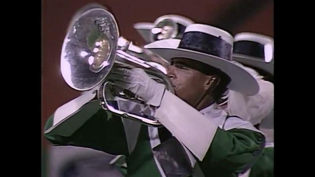 A musician in a green uniform and large white hat plays a mellophone, with other drum corps members visible in the background. The scene captures a moment during a performance.