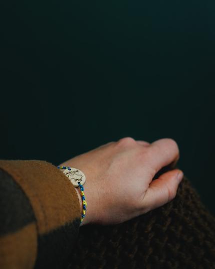 A person’s wrist wearing a handmade bracelet in blue and yellow with a wooden centerpiece engraved with the outline of Ukraine and the word “UKRAINE.” The person is dressed in a brown coat and holding a textured brown fabric.