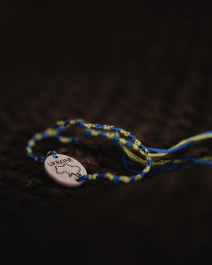 A close-up of a handmade blue and yellow string bracelet with a wooden centerpiece engraved with the outline of Ukraine and the word “UKRAINE,” lying on a dark textured surface.