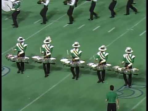 A group of drummers in green and white uniforms perform on a football field. They play multi tenor drums while a few performers move in the background. The scene captures a dynamic moment of a drum corps performance.