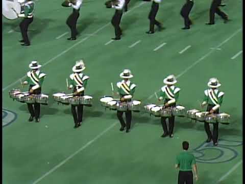 A group of drummers in green and white uniforms perform on a football field. They play multi tenor drums while a few performers move in the background. The scene captures a dynamic moment of a drum corps performance.