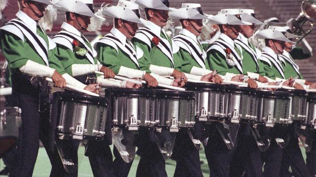 A group of drummers in green and white uniforms perform in a line, each holding drumsticks. They are playing snare drums. The setting is on a football field.