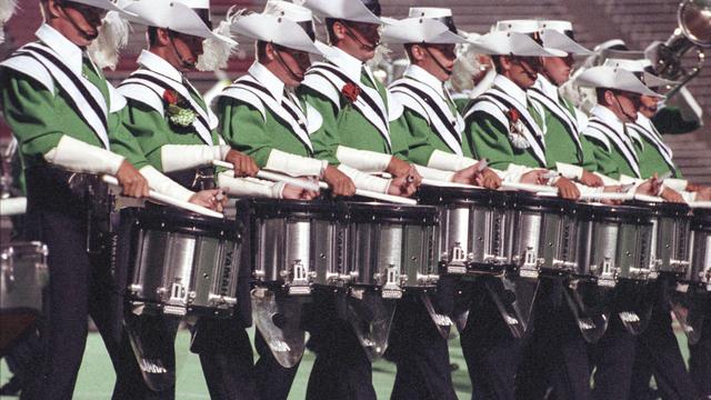 A group of drummers in green and white uniforms perform in a line, each holding drumsticks. They are playing snare drums. The setting is on a football field.