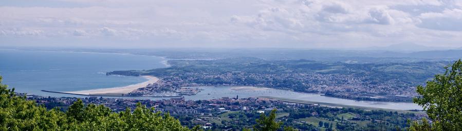 A panoramic view of a coastal landscape, featuring a sandy beach, a river leading to the ocean, and a town with various buildings and greenery in the foreground. The sky is partially cloudy, and the horizon features rolling hills in the distance.