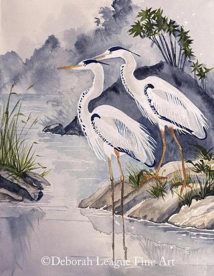 Elegant Watercolor Art Print of two herons at Water's Edge.  Two tropical birds with striking blue and white plumage and long legs stand near the edge of a body of water, surrounded by rocks and sparse vegetation. The background features a misty, wooded landscape, giving a sense of tranquility to the scene.