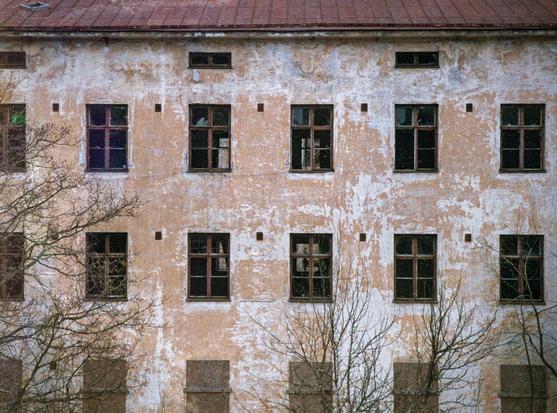 A close-up of a multi-storey, abandoned building. A lot of dark, broken windows. Shabby walls with shabby orange paint from under which light gray concrete is visible. Leafless shrubs on the edge and at the bottom of the image.