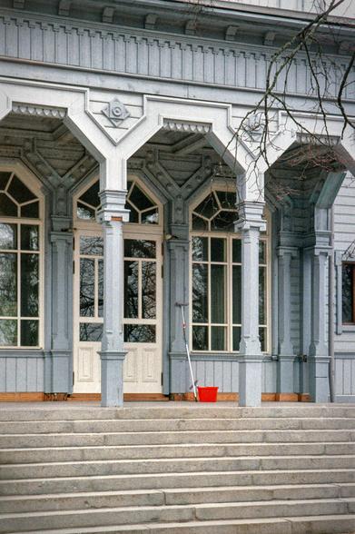 An antique, wooden house of gray-blue color. Large windows with white frames. Tall staircase and wooden columns with ornamentation. A red bucket and a mop stand against the wall of the house.