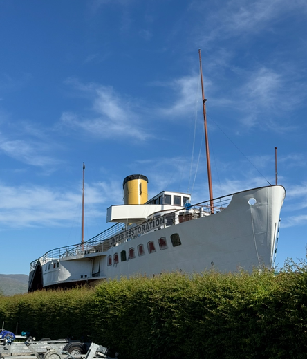 1950s disused paddle steamer raised on a slipway for renovation works. It’s seen better days but looks majestic in plain white paint with yellow raking funnel, all under a clear blue sky.