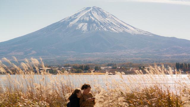 The image features a scenic view of a snow-capped Mount Fuji, in the background. In the foreground, two people are standing near a body of water and tall grass. The scene suggests a peaceful, natural setting.