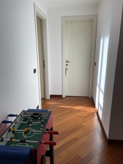 L'immagine mostra un corridoio con pavimento in legno di colore marrone chiaro, che riflette la luce naturale proveniente da una finestra a destra. Le pareti sono dipinte di bianco, creando un'atmosfera luminosa e spaziosa. In primo piano, a sinistra, si trova un tavolo da calcio, con manopole rosse e blu e figure di giocatori in giallo e blu, pronto per un gioco. In fondo al corridoio, si vede una porta bianca con una maniglia e una tassella, che sembra essere chiusa. A sinistra, c'è un'altra porta aperta che conduce a un'altra stanza. L'ambiente è semplice e minimalista, con un'attenzione particolare alla pulizia e all'ordine.