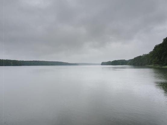 The image shows a calm, expansive body of water, possibly a lake or river, surrounded by dense green forest. The sky is overcast with gray clouds, suggesting a cloudy or rainy day. The water surface reflects the gray tones of the sky.