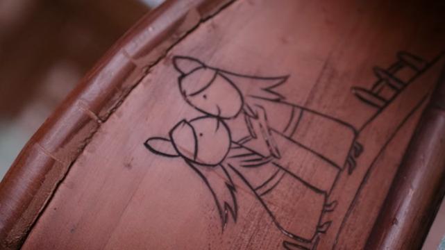 Close up on a piece of wooden furniture into which a scene has been carved. The scene features a couple standing on a road, little fences carved in beside the road beyond them in either direction. The figures are essentially identical aside from the one on the left being a little shorter and are facing each other, sharing a happy kiss while holding hands.