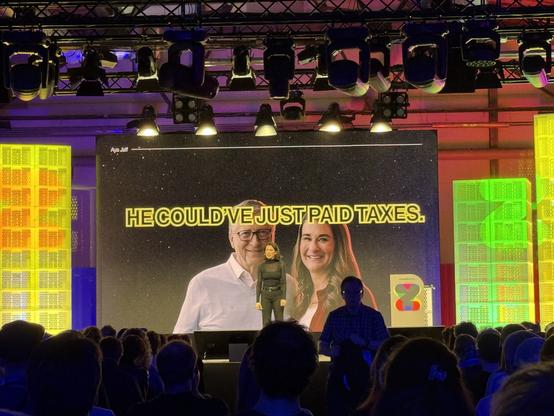 A speaker stands on stage in front of a large screen displaying a notable quote: "HE COULD'VE JUST PAID TAXES." The screen features images of two individuals, one is Bill Gates and the audience is visible in the foreground. The event appears to focus.