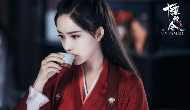 Close in shot of Wen Qing in vivid red robes over a white underlayer, with a cup raised cup raised to her mouth from which she sips.