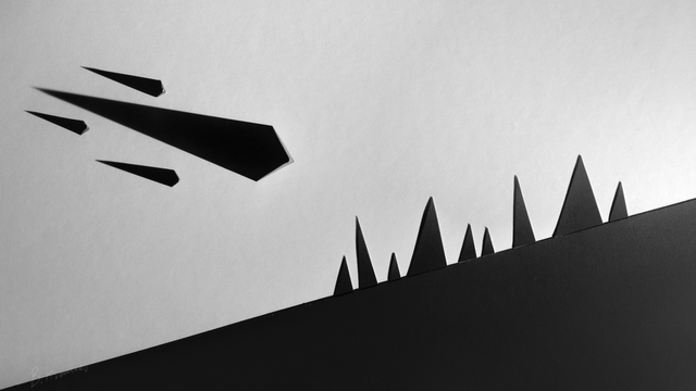 A high-contrast black and white image of a slanted black surface portraying a hillside, with smaller black triangles clustered together like a forest or city buildings. Additional black poster-board triangles have been folded partially and stood up, and a harsh spotlight makes them cast a long shadow, making them look like meteors with a long trailing shadow.