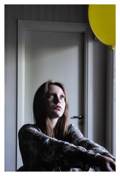 En stående bild på en flicka som gör ett bagger-slag mot en gul ballong. Hon har en mönstrad blues och tittar mot ballongen som är uppe till höger. I bakgrunden en vit dörr. Ljuset kommer från fönster till höger.