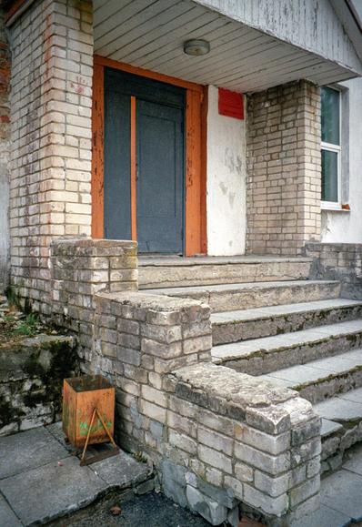 The photo shows the entrance to a white, brick office building. The area around the door is painted with orange paint. Next to the entrance is an orange urn.