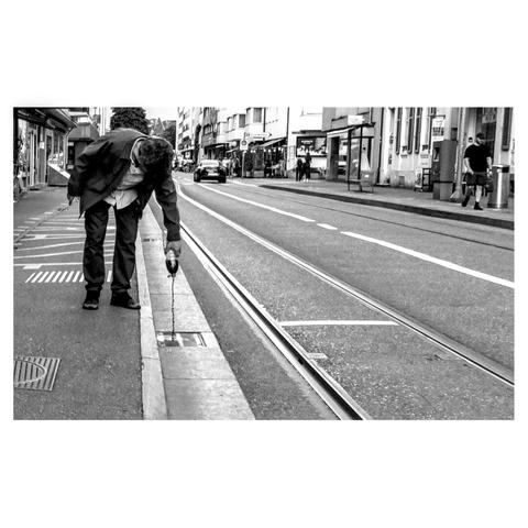 A black and white phtograph with pushed contrast to highlight diagnoal lines depicting a man bending over to pour a bottle of lemonade (coke or similar) down the gully/drain of a street. There are tram tracks on the street.