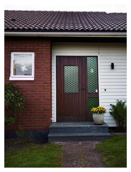 En stående bild på en brun trädörr med grönt mönstrat glas. På dörrens högra sida finns två sektioner av likadant glas. Väggen är av vitt trä till höger, rött tegel till vänster. Taket har svarta takpannor. Stenplattor leder fram till dörren, gräsmatta på båda sidor. En grå stenkruka med gula blommor står på den blygråa trappan.