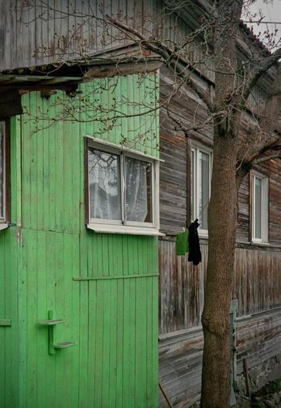 The photo shows the wall of an old, wooden house. Part of the wall is painted in green color. A rope is stretched between the wall and the village. The laundry is drying on the rope, part of which is green.