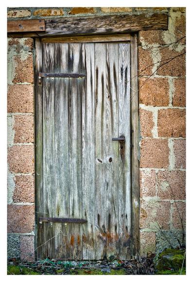 The picture shows an old wooden door built into a wall of rough, reddish-brown bricks. The door is made of vertical, weather-beaten wooden planks that are discoloured grey-brown and have dark stains and slight cracks in places. The door handle is plain and made of metal. A solid, weathered wooden beam runs above the door. Some moss and dry grass grow on the ground in front of the door. The entire scene looks old, abandoned and marked by the weather.