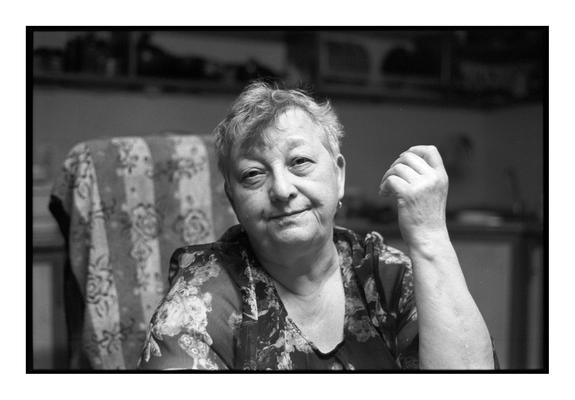 A portrait of an elderly woman sitting in a kitchen, looking directly at the camera with a calm and thoughtful expression. Shot on black and white film (Ilford Pan 400) with a Minolta Dynax 7