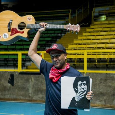 Tom Morello con gorra roja y pañuelo rojo al cuello, sostiene una guitarra acústica sobre su cabeza con su mano izquierda. Con su mano derecha sostiene una imagen en blanco y negro de Victor Jara fumando. El fondo es una cancha deportiva con gradas amarillas.